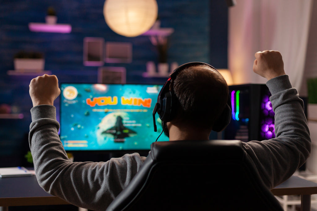 Excited player with headphones making winner gesture while playing games on computer at gaming home studio.